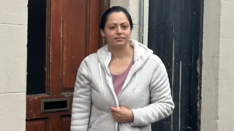 Mika Kolev is a young woman with dark hair which is pulled back into a ponytail.
She is wearing a grey cardigan over a pink top.
She is standing at her front door which was damaged on Tuesday night.
