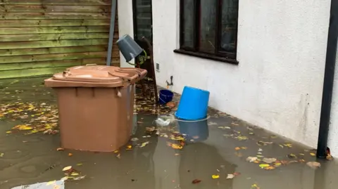 Steve Pelham Dirty water half way up the wall of a house and a brown wheelie bin. There is a bucket and lots of brown leaves floating in the water.