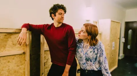 Crossed Wires Greg James is tall with a red long-sleeved shirt and medium-length curly brown hair. He is looking over his shoulder to Alice, who has ginger hair and a floral blue and white blouse, who looks up at him.