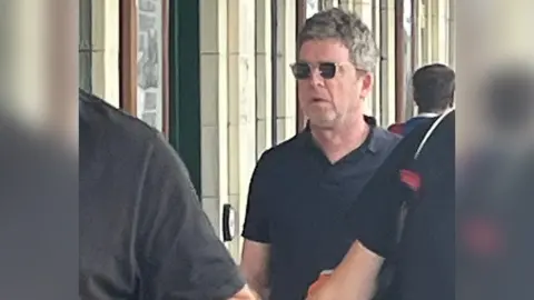 Noel Gallagher on a platform at Cardiff Central Railway Station. He is wearing a black, collared T-shirt and black sunglasses with clear frames. There are three unidentifiable people around him as he walks through a crowd and white brick walls are behind him