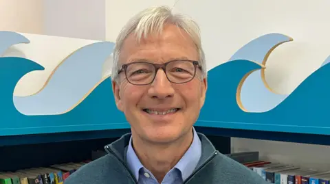 Nick stands in front of a bookshelf filled with colorful books in what appears to be a library or reading area. The shelving has a decorative wave design in blue and white.