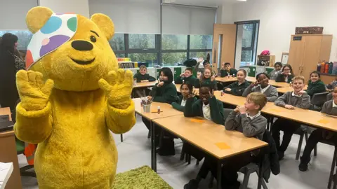 Yellow Pudsey bear waving at the camera as children sit behind at desks and smile at the camera and give peace signs.