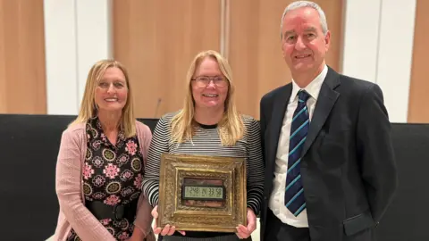 Campaigners Anne, Carolyn Basing and Nigel Taylor smiling at the camera in front of the council chamber's lectern. Anne and Carolyn both have straight blonde hair and blue eyes. Anne is wearing a black belted dress with green and pink flowers and a pink cardigan. Carolyn is wearing a black and white striped bloused as she holds a gold-frames electronic clock showing there were 248 days, 14 hours, 33 minutes and 58 seconds until the glass centre's closure. Nigel has short white hair and is wearing a black suit with a white shirt and a blue tie.