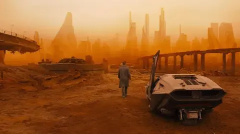 Warner Bros Gambar diam dari film Blade Runner 2049, yang menunjukkan kendaraan di sebelah kanan gambar dan seseorang berjalan menuju kota yang tampak distopia dengan warna oranye.