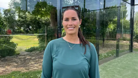 Kate, wearing a turquoise top, looks at the camera. She has dark brown shoulder-length hair which is tied up, with sunglasses on the top of her head. She has a silver necklace on, and is stood in a padel court with the glass wall behind her.