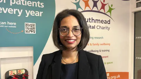 Dr Kathreena Kurian standing in front of Southmead Hospital Charity logo.