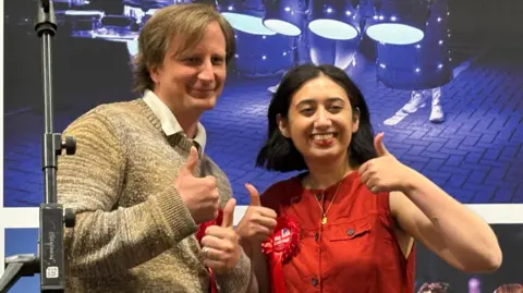 Louis Brand/BBC Shanika Mahendran, standing in front of a banner, with short bobbed hair, wearing a red top, with a rosette on her right side, holding up her thumbs, smiling. She is next to a man, with short hair, wearing a white shirt and jumper. They are by a microphone stand. 