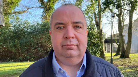 Brian Tierney in a light blue shirt and navy jacket standing near trees.