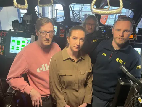 Four people stand closely together inside an RNLI lifeboat, surrounded by navigation screens and control panels. On the right, stand two RNLI crew members in navy uniforms with a woman in a khaki-coloured shirt and a man in a pink sweatshirt standing to their left.