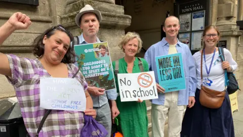 A group of parents holding signs against smartphones in schools. They are smiling for the camera. One woman has her fist up in the air.