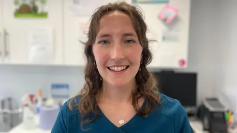 A close-up of a woman with brown hair. She is wearing blue scrubs and smiling as she stands in veterinary surgery.