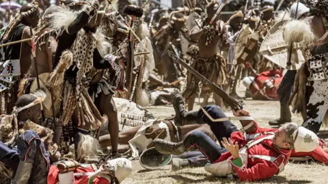 AFP/Getty Images A enactment in 2023 of the Battle of Isandlwana with Zulu warriors with shields and spears bearing down on British soldiers with guns in red jackets and white helmets, several of whom have fallen to the ground. 