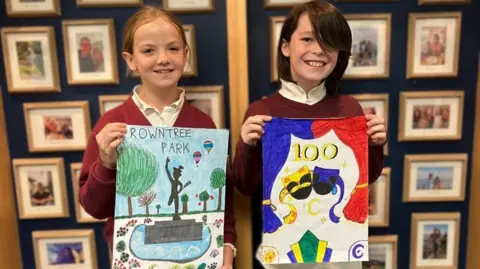 A girl and boy stand holding pieces of art - one reads "Rowntree Park" and has a drawing of a statue, trees and hot air balloons - the other reads "100" and has pictures of theatre curtains and masks. They are looking at the camera and smiling.