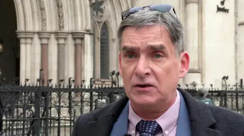 Gary Webster gives a TV interview outside the High Court in London. He has parted grey hair with sunglasses over his head and wears a jacket, shirt and tie with a dark coat.