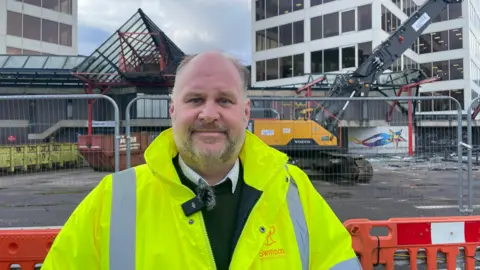 Man in high vis jacket standing in front of fence, a digger and office buildings.