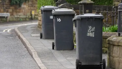 Three black wheelie bins stand on a pavement positioned in front of a fenced area with stone pillars.