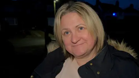 Nicola Daniels stands outside in a road in the city. It is night-time and there is a light behind her. She is wearing a coat with a fluffy hood.