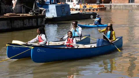 Gloucester City Council A family sit in two blue canoes padding through the docks. There are two children in orange/red life jackets, and their parents are sat behind them in blue life jackets.