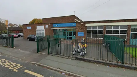 Google A Google street view image of the front of a one-storey school building. A green fence is at the front, with some bikes in a courtyard in front of the front doors. A car park is visible to the left of the building.