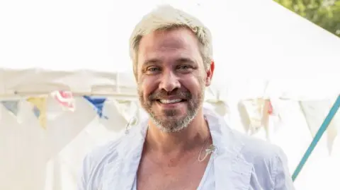 Getty Images Will Young, a man with light blonde hair and grey stubble and wearing a white shirt, smiling at the camera. There's a white marquee and some bunting behind him and it's a sunny day.