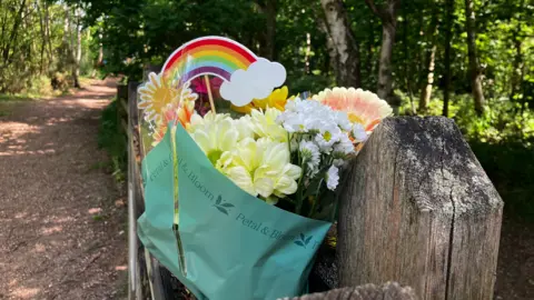 Jessica Lane/BBC Un ramo de flores de colores atado a una verja. Detrás hay un sendero polvoriento y un bosque.
