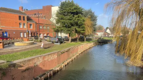 A view of the town centre taken from the vantage point of a bridge over the river with branches of a willow tree overhanging the water to the right of the picture. It is a sunny day and to the left, on the river bank, you can see a wooden seating area in front of red brick shop buildings.