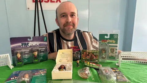 Craig crouches down poses behind a table on which there are various models from his collection. He is balding with a black and grey stubbly beard and is wearing a white, brown and black vertically-striped t-shirt.