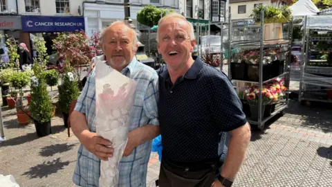 Kevin Shoesmith/BBC Two men, including Andrew Dennis, on a market stall. The other man is holding a bouquet of flowers