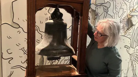 Leeds Museums and Galleries A woman with white hair, wearing a green top and black-rimmed glasses rings a large bell in a wooden frame.