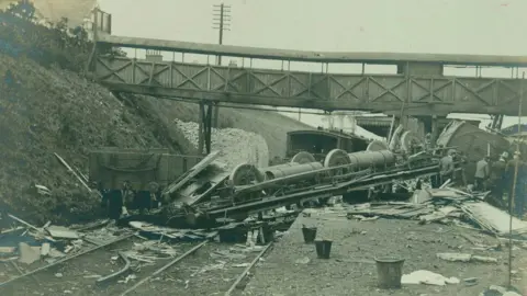 Fred Spalding A black and white photograph showing the aftermath of the Witham railway crash. A carriage is lying upside down across the platform, its wheels and metal framework exposed. The platform is covered in debris. There is a bridge over the railway tracks, and people are stood on the platform.