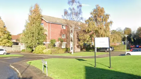 A Google Street image of a red-bricked building on a residential road. There is a white sign on a grass mound out the front which says "care home" on it.