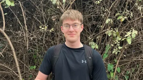 A portrait image of local student George, 21, stood on the pavement of the road which crosses Lady Bay Bridge. There is a bush behind him in the background.