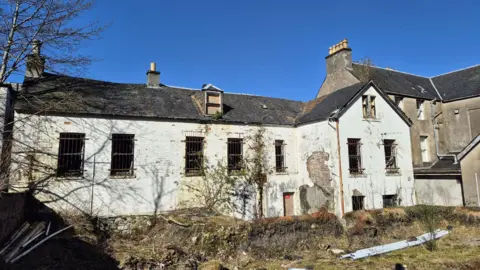 Ian Peter MacDonald A large derelict building with crumbling white washed render. There are boarded up windows and the area next to the building looks overgrown and abandoned.
