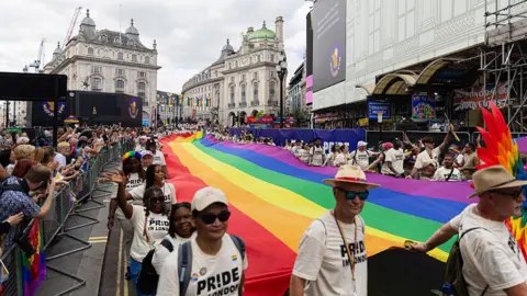 Participants wearing matching "Pride in London" T-shirts carry a giant, long rainbow flag down a street lined with spectators in London's Piccadilly Circus during a Pride parade.