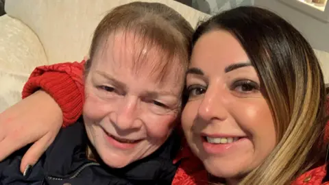 Nicola Hughes is a selfie of Nicola Hughes (right), which has her arm around her mother Janet Richie. They are sitting on a cream couch and smiling on the camera.