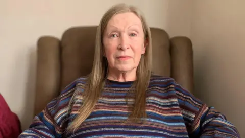 BBC A woman with long, light brown/blonde hair in a striped jumper sitting on a brown leather chair