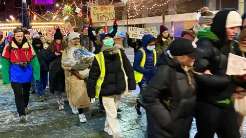 University of Sheffield Students' Union A crowd including men and women gathered in front of buildings lit by Christmas lights. Some are holding banners which read "The only cause of rape is rapists" and "Harassment is not a compliment".