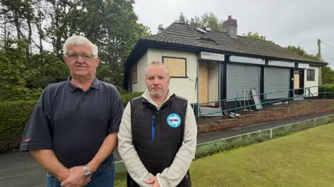 Barrie Bulch is wearing a blue polo shirt and is standing next to Councillor Paul Donaghy, who is wearing a black gilet with a blue Reform UK logo. They are standing on a bowling green in front of the burnt-out club house behind them.