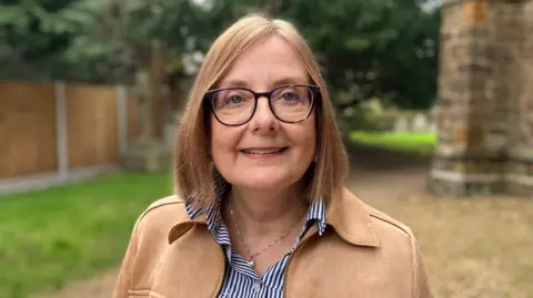Caroline Barazesh, a woman who is standing next to a church. She is looking directly at the camera and smiling. She is wearing a pair of glasses over her eyes and is wearing a blue and white striped shirt and brown suede jacket.