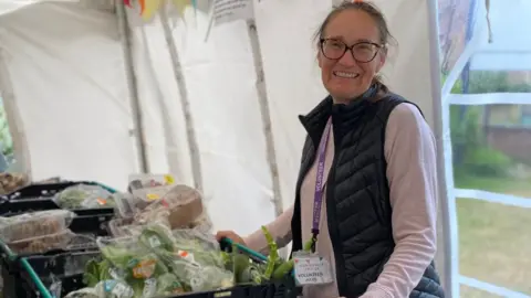 Jules Hopkins A woman with glasses wearing a black gillet and purple shirt smiling at the camera. She is standing in a marquee in front of crates of food.