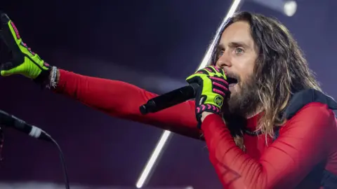 Jared Leto performs on stage in a red mesh top. He is singing into a microphone and wearing lime green gloves. 