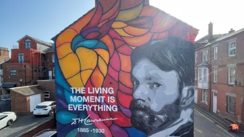 A mural on the side of a building on a street in Eastwood. There is a colourful phoenix in a stained-glass window style with a painted portrait on the right of DH Lawrence in black and white. On the left, there is a quote by DH Lawrence saying "The Living Moment is Everything", with his years of birth and death - 1885 and 1930 - underneath