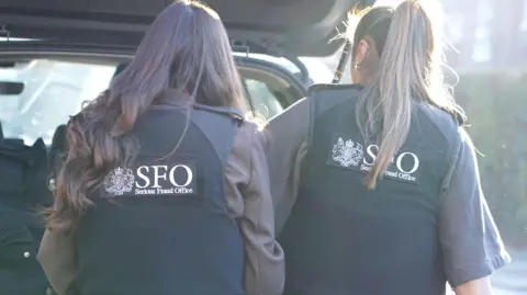SFO Two Serious Fraud Office officers are seen from behind wearing SFO-branded vests, standing at the open boot of a car in daylight.