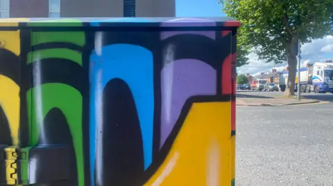 Utility box with crayons painted on it. A road is visible behind.