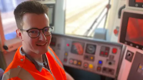 Liam smiling at the camera, wearing an orange high-vis jacket, sitting in the front of a train, beside the control panel 