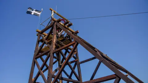 King Edward Mine/ Toby Weller A large wooden tower with supports to its left and a wheel at the top is flying the black and white Cornish flag. 