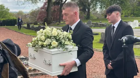 A man in a black suit carries a small white coffin with a large bouquet of white flowers on its lid.