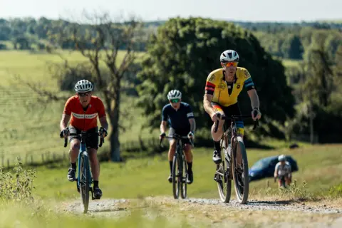 Red On Sports Four cyclings head up a hill on a gravel path through lush, green countryside