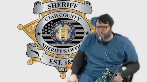 Fox 13 A video link image of Nicholas Rossi in blue prisoner clothes, sitting in a wheelchair in front of an image of the Utah County Sheriff's Office bade. He is wearing an oxygen mask and holding an oxygen bottle in his left hand