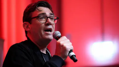 Andy Burnham, a man with short dark hair and black-rimmed glasses, speaks into a microphone. He is wearing a dark zip up top and against a red backdrop